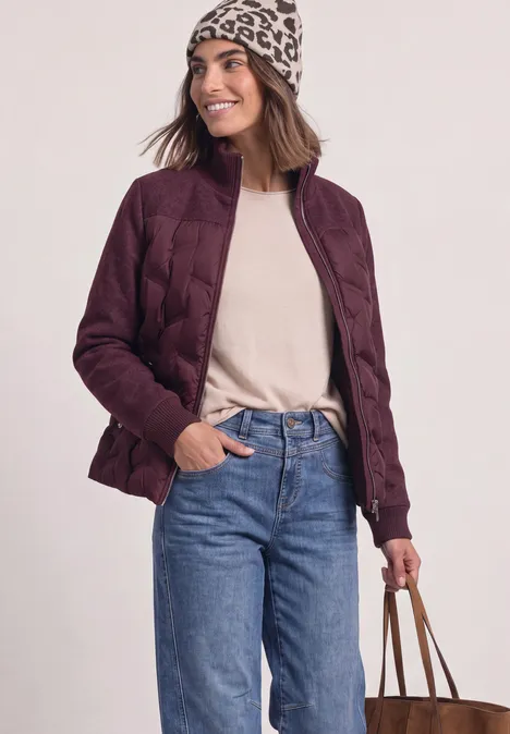 Frau trägt burgunderrote Steppjacke, Jeans und Beanie, hält eine Tasche.