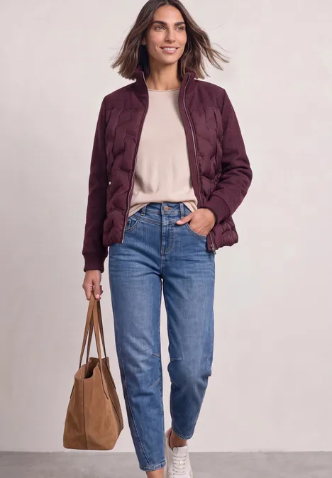 Frau trägt eine bordeauxrote Steppjacke, beiges Shirt, blaue Jeans und eine braune Tasche.