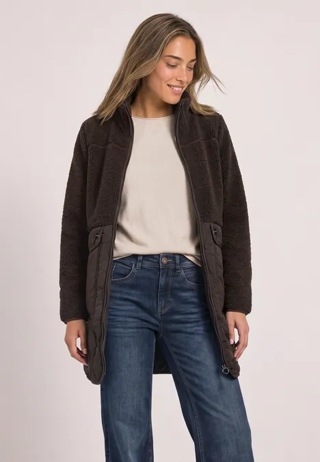 Frau in braunem Mantel, weißem Shirt und Jeans, lächelnd, blickt nach links.