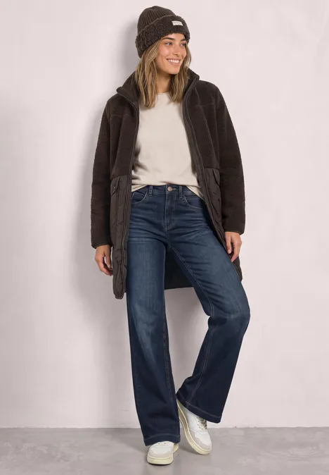 Fröhliche Frau in Beanie, weißem Shirt, braunem Mantel, weiten Jeans und weißen Sneakern vor neutralem Hintergrund.