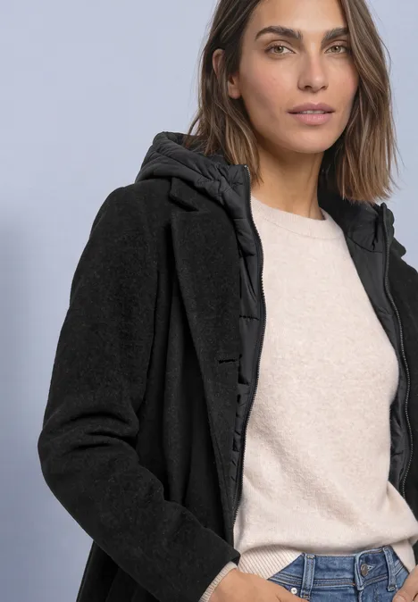 Manteau à col à revers et capuche intégrée Black Femme en manteau à capuche noir doublé, pull beige et jean, posant devant un fond simple.