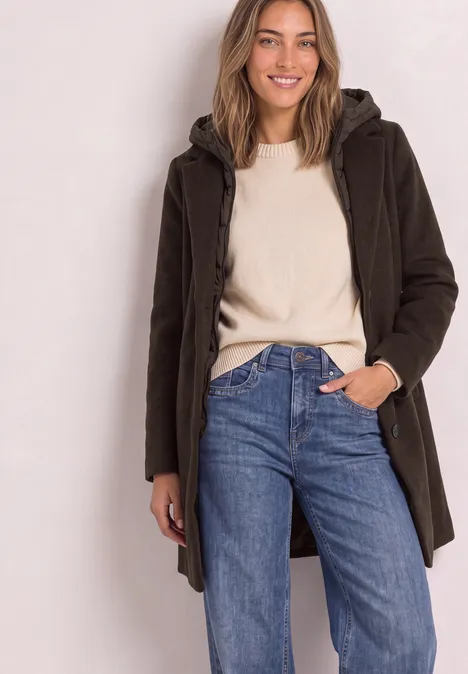 Junge Frau trägt braunen Parka über hellem Pullover und blauen Jeans, posiert lässig.