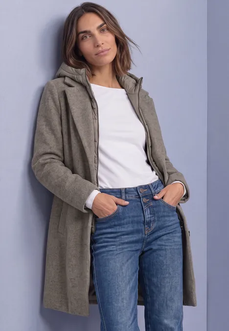 Femme en manteau vert-gris, t-shirt blanc et jeans, mains dans les poches contre un mur.