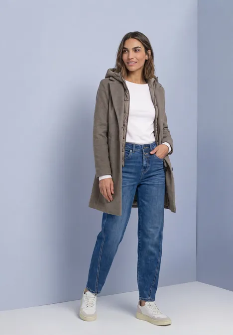 Femme en manteau kaki à capuche, top blanc, jean bleu taille haute et baskets blanches.
