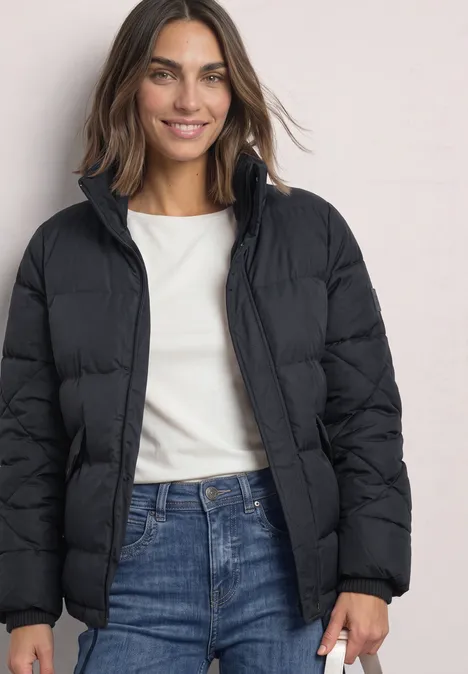 Steppjacke mit Stehkragen und Tunnelzug Obsidian Blue Frau in schwarzer Steppjacke, weißem Shirt und Jeans steht lächelnd vor neutralem Hintergrund.