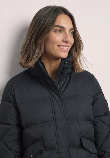 Steppjacke mit Stehkragen und Tunnelzug Obsidian Blue Lächelnde Frau in einem schwarzen Daunenmantel mit dunklen Haaren, leicht seitlich zur Kamera blickend.
