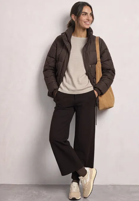 Vrouw leunt tegen muur, bruine donzen jas, licht truitje, wijde donkere broek, sneakers en schoudertas.
