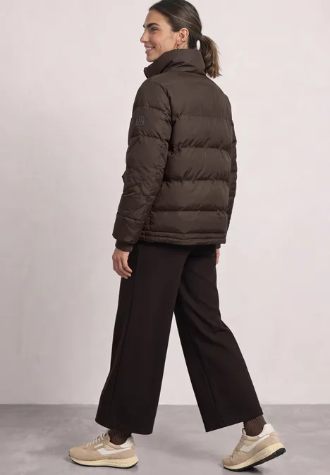 Vrouw van achteren met een bruine gewatteerde jas, brede zwarte broek en beige sneakers.