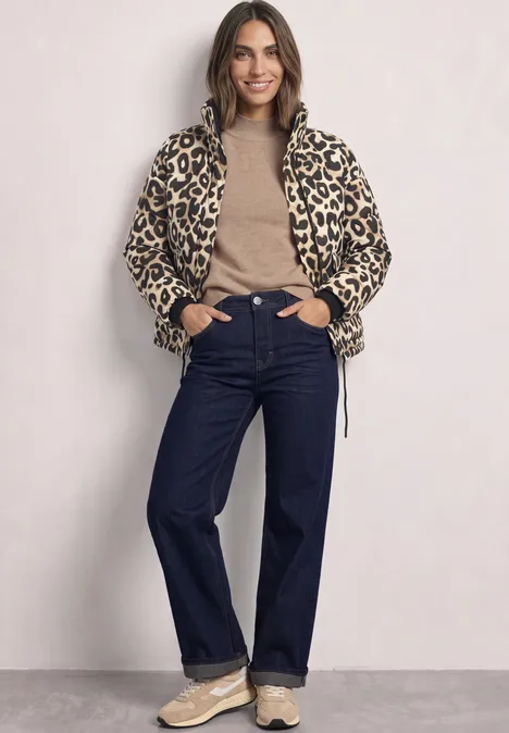 Vrouw met luipaardprintjas, beige trui, donkere jeans en sneakers, lachend voor effen achtergrond.
