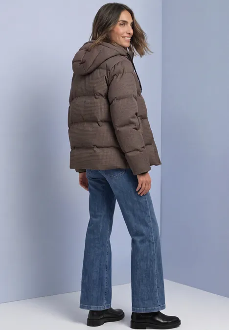 Frau in brauner Steppjacke mit Kapuze, Jeans und schwarzen Schuhen, schaut lächelnd über die Schulter.