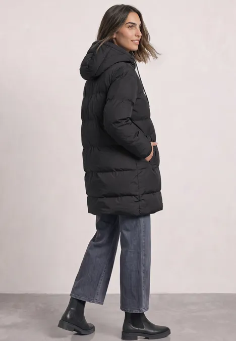 Frau von der Seite im schwarzen Steppmantel mit Kapuze, blauen Jeans und schwarzen Stiefeln.