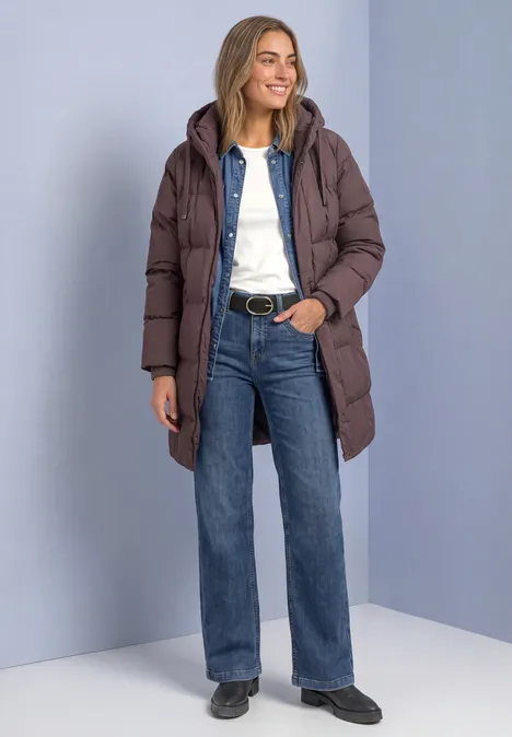 Frau in langer brauner Steppjacke, weißem Oberteil, weiten Jeans und schwarzen Schuhen vor grauem Hintergrund.