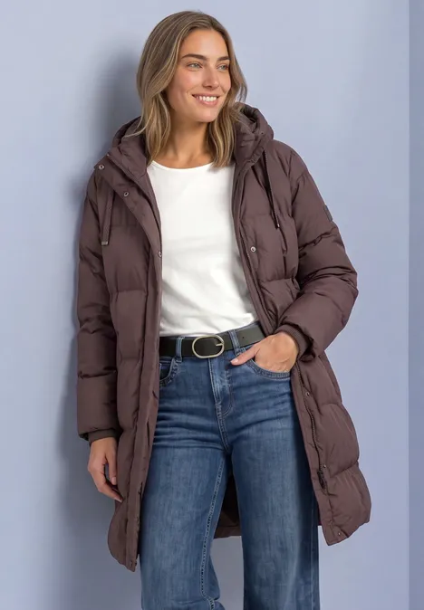 Frau trägt lange braune Steppjacke, weiße Bluse, blaue Jeans, lächelt vor neutralem Hintergrund.