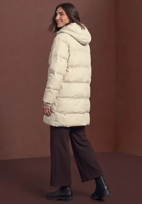 Frau von hinten in cremefarbener Steppjacke mit Kapuze, braunen Hosen und schwarzen Stiefeletten vor braunem Hintergrund
