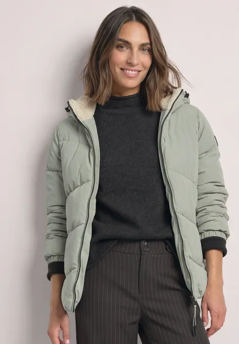 Frau mit grüner Steppjacke und schwarzem Pullover lächelt vor hellem Hintergrund.
