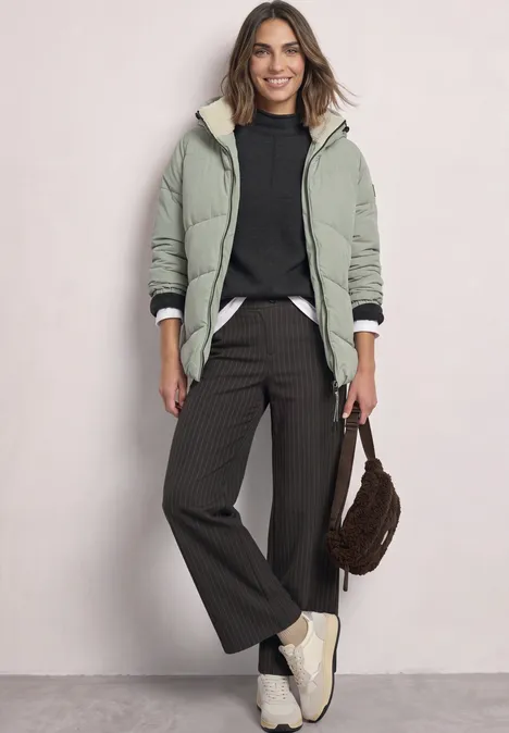 Frau in grüner Jacke mit Teddyfutter, schwarzem Pullover, weiter Hose und weißen Sneakern, braune Tasche.
