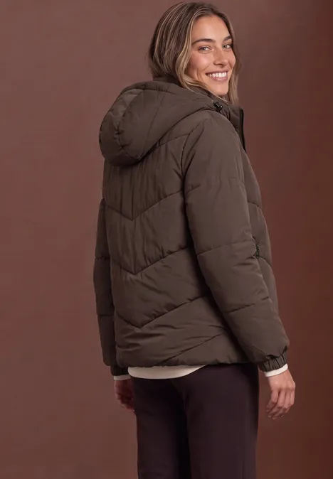 Eine Frau trägt eine braune Steppjacke mit Kapuze und lächelt über die Schulter.