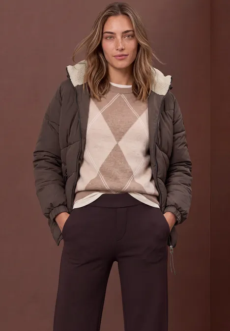 Frau mit brauner Steppjacke und beigem Argyle-Pullover, dunkle Hose, Hände in den Taschen.