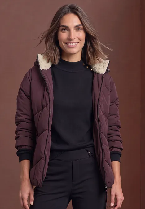 Frau in bordeauxroter Steppjacke mit Kapuze, schwarzem Oberteil und Hose vor braunem Hintergrund.