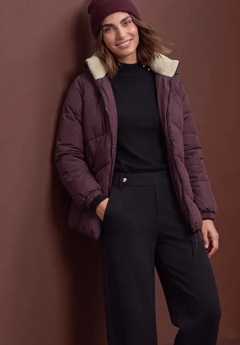 Frau in lila Winterjacke mit Fellkragen, Mütze, schwarzem Pullover und Hose, lächelnd vor braunem Hintergrund