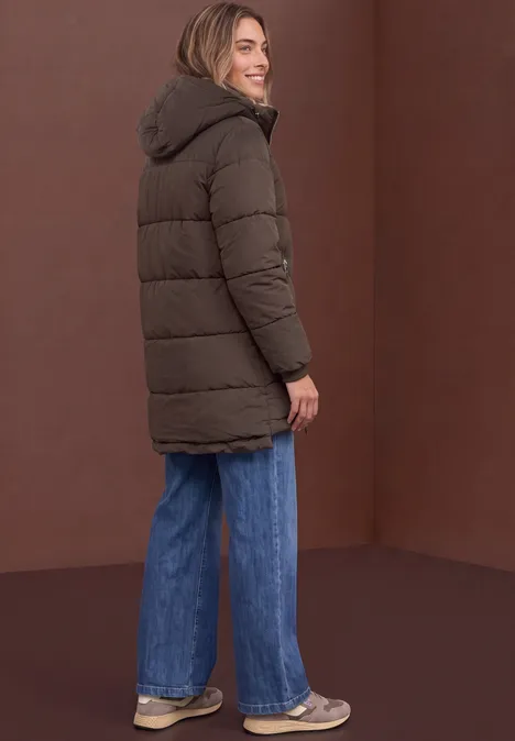 Vrouw van opzij in bruine gewatteerde jas met capuchon, blauwe wijde jeans en sneakers voor een bruine achtergrond.