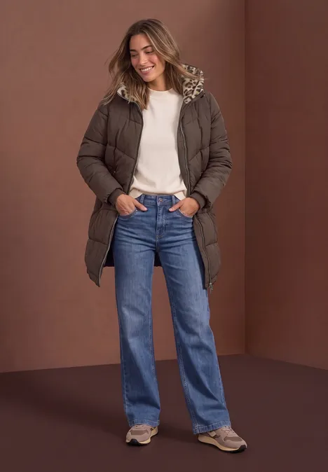 Vrouw in bruin gewatteerd jasje, wit sweater en blauwe jeans, glimlachend voor een bruine achtergrond.