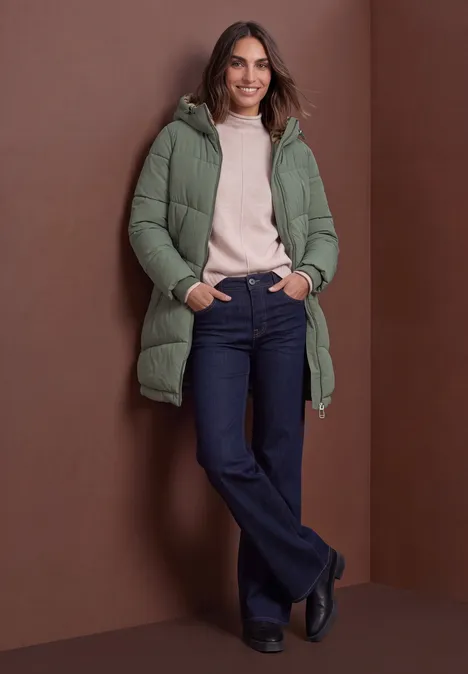 Frau vor braunem Hintergrund, in grünem Steppmantel mit Kapuze, rosa Pullover, Jeans und schwarzen Schuhen, lächelt.