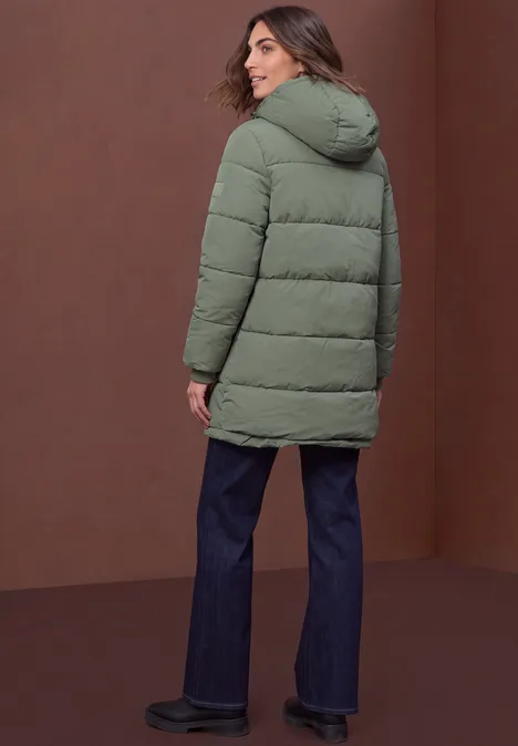 Frau in grüner Steppjacke, blauen Jeans und schwarzen Schuhen, steht vor braunem Hintergrund und blickt zurück.