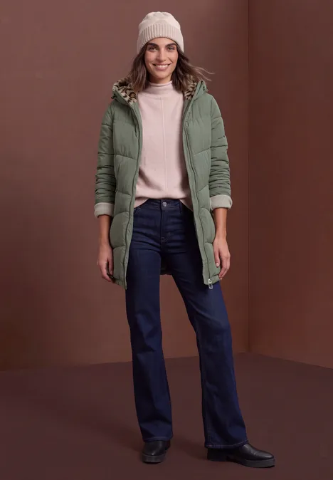Frau in grünem Wintermantel mit Kapuze, heller Mütze, Pullover, dunkler Jeans und schwarzen Stiefeln.