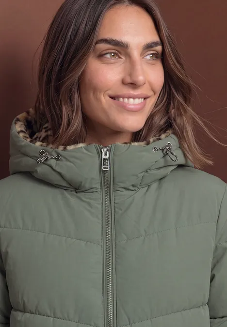 Eine lächelnde Frau trägt eine grüne, gesteppte Jacke mit Kapuze vor braunem Hintergrund.
