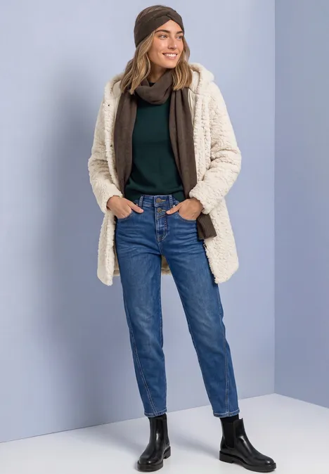 Teddymantel mit gefüttertem Inlay smooth stone sand Frau in Jeans, schwarzem Rollkragenpullover, Beanie, cremefarbener Teddyjacke und schwarzen Boots lächelt.