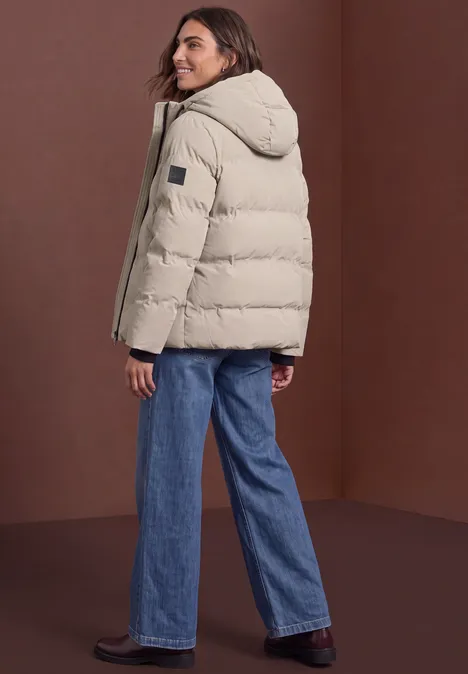 Frau in beige Daunenjacke mit Kapuze und blauen Jeans, seitlich stehend, blickt über die Schulter
