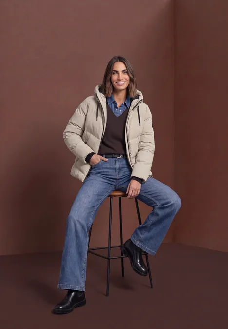 Frau in beiger Steppjacke, Hemd und Jeans sitzt lässig auf einem Hocker vor braunem Hintergrund.