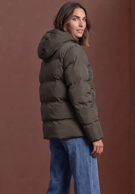 Pufferjacke mit Kapuze auburn brown Frau in dunkelgrüner Steppjacke und blauen Jeans blickt über die Schulter.