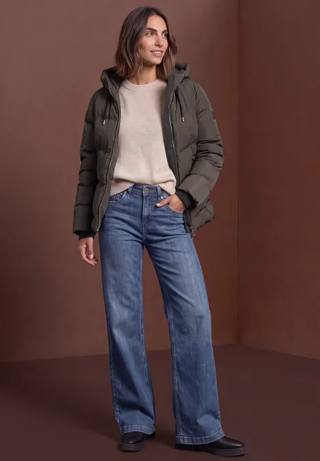 Pufferjacke mit Kapuze auburn brown Frau trägt grüne Steppjacke, beigen Pullover und weite blaue Jeans vor braunem Hintergrund.