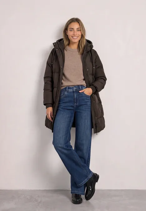 Lange Jacke mit Kapuze und Steppmuster auburn brown Junge Frau in braunem Steppmantel, beigem Pullover und Jeans vor neutralem Hintergrund, lächelnd.