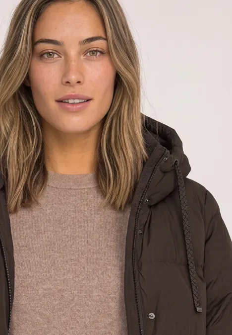Lange Jacke mit Kapuze und Steppmuster auburn brown Junge Frau mit offenen braunen Haaren trägt einen braunen Steppmantel und Pullover, vor hellem Hintergrund.