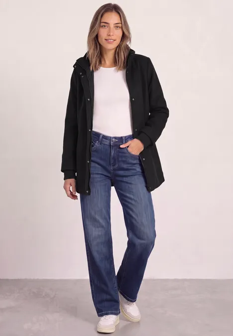 Frau in schwarzem Mantel, weißem Top, blauen Jeans und weißen Sneakers steht vor hellem Hintergrund.