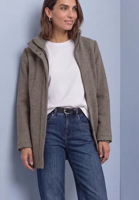 Kurzer Mantel mit Strickdetails Muddy Mocha Melange Frau in weißem T-Shirt, blauen Jeans und grauer Kapuzenjacke steht vor einfarbigem Hintergrund.