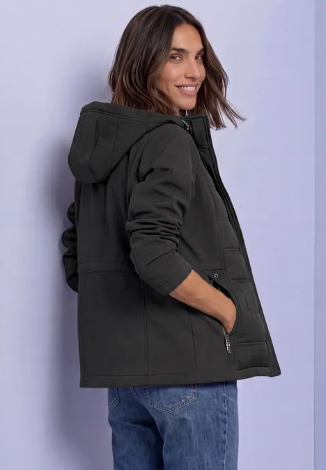 Veste softshell à capuche avec détails matelassés Black Frau in schwarzer Kapuzenjacke, lächelnd seitlich posierend vor hellem Hintergrund.