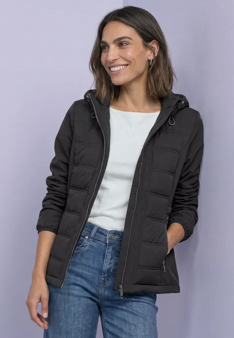 Veste softshell à capuche avec détails matelassés Black Frau in schwarzer Steppjacke mit Kapuze, weißem Shirt und blauen Jeans, lächelnd.