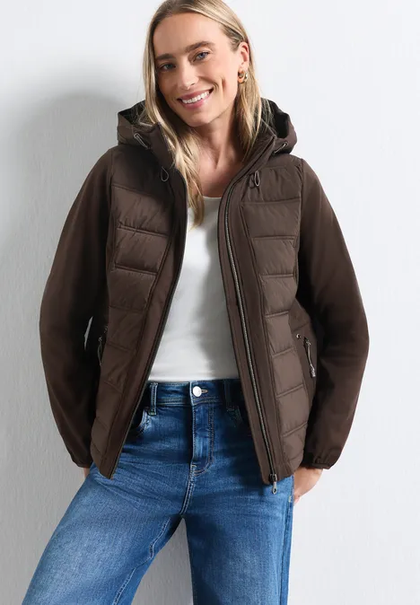 Frau in brauner Kapuzenjacke, weißem Top und blauen Jeans vor neutralem Hintergrund, lächelnd
