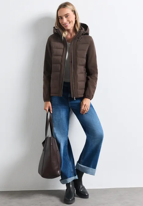 Frau in brauner Steppjacke, weitem Jeans, schwarzen Stiefeletten und Tasche vor weißer Wand.