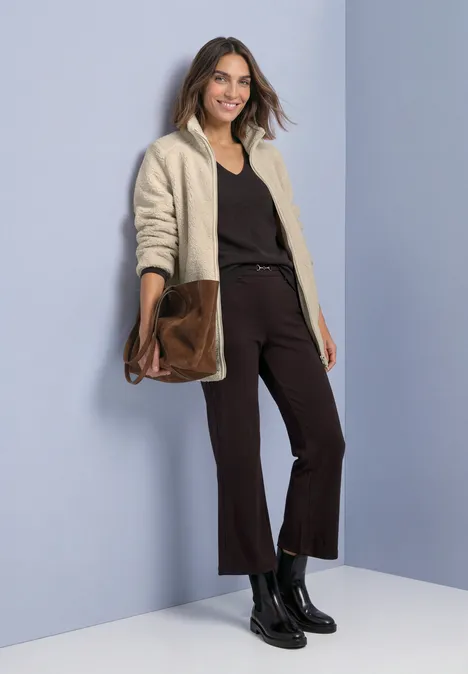 Frau lehnt an blauer Wand, trägt beigen Cardigan, schwarzen Pullover, schwarze Hose und Stiefel, hält braune Tasche.