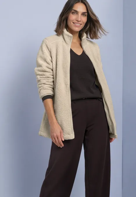 Eine Frau trägt eine beige Teddyjacke, schwarzes Top und Hose, sie lächelt vor hellem Hintergrund.