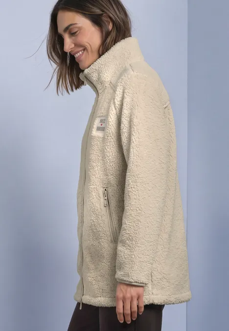 Eine Frau trägt lächelnd eine hellbeige Teddy-Fleecejacke mit Stehkragen vor hellem Hintergrund.