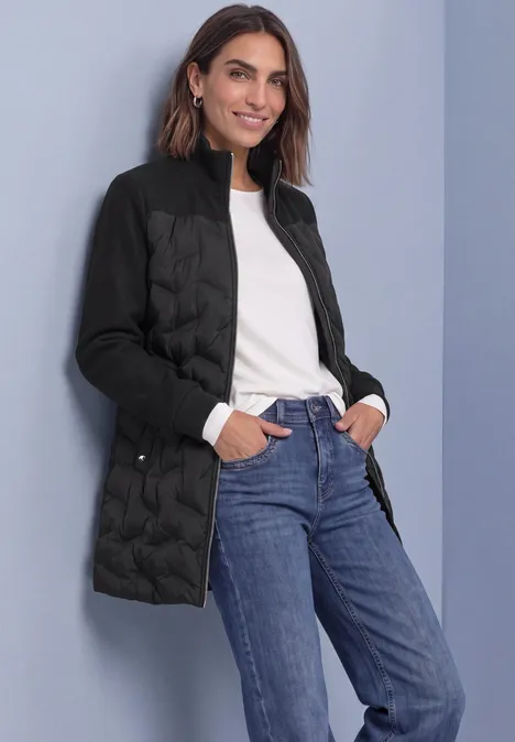 Strukturmix Jacke mit geklebter Steppung Black Frau in schwarzer Steppjacke, weißem Oberteil und blauen Jeans, lächelnd mit Händen in den Taschen vor grauem Hintergrund.