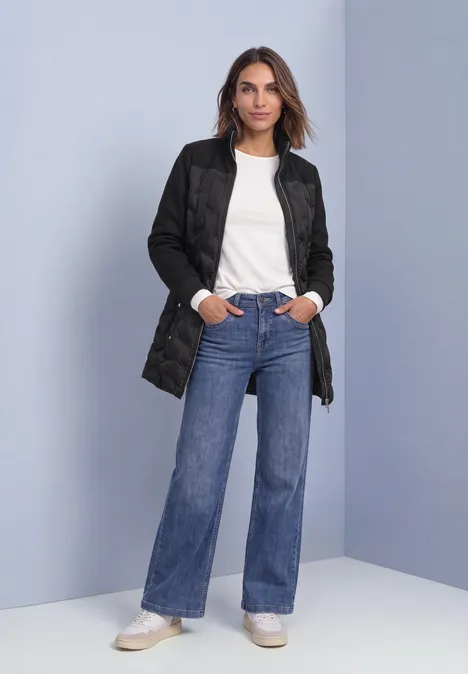 Strukturmix Jacke mit geklebter Steppung Black Frau in langer gesteppter schwarzer Jacke, weißem T-Shirt, weiten Jeans und weißen Sneakern vor schlichtem Hintergrund.