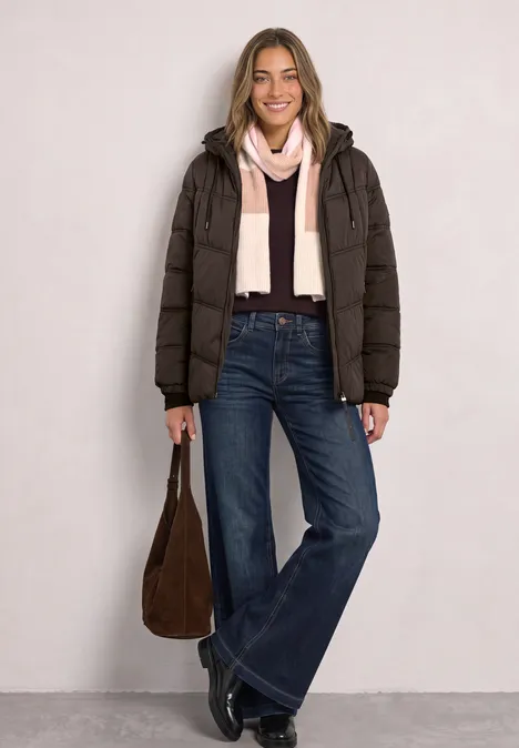Frau im Steppmantel mit Jeans und rosa Schal, hält eine braune Tasche und lächelt.