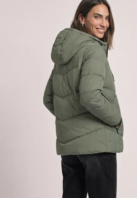 Frau in grüner Steppjacke mit Kapuze, lächelnd seitlich zur Kamera gewandt.